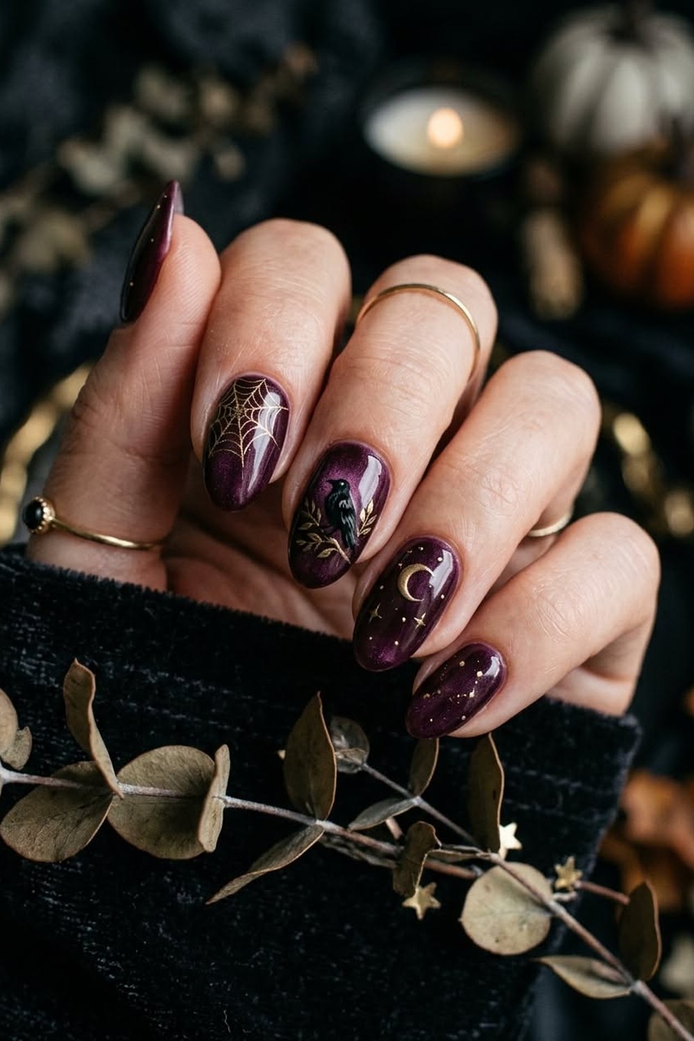 Close-up of a hand with medium almond nails in glossy deep plum gel with a soft shimmer, a gold spiderweb on the index nail, a small black raven on a gold branch, a gold crescent moon with tiny stars and scattered gold dots, held over a black velvet sleeve with dried eucalyptus