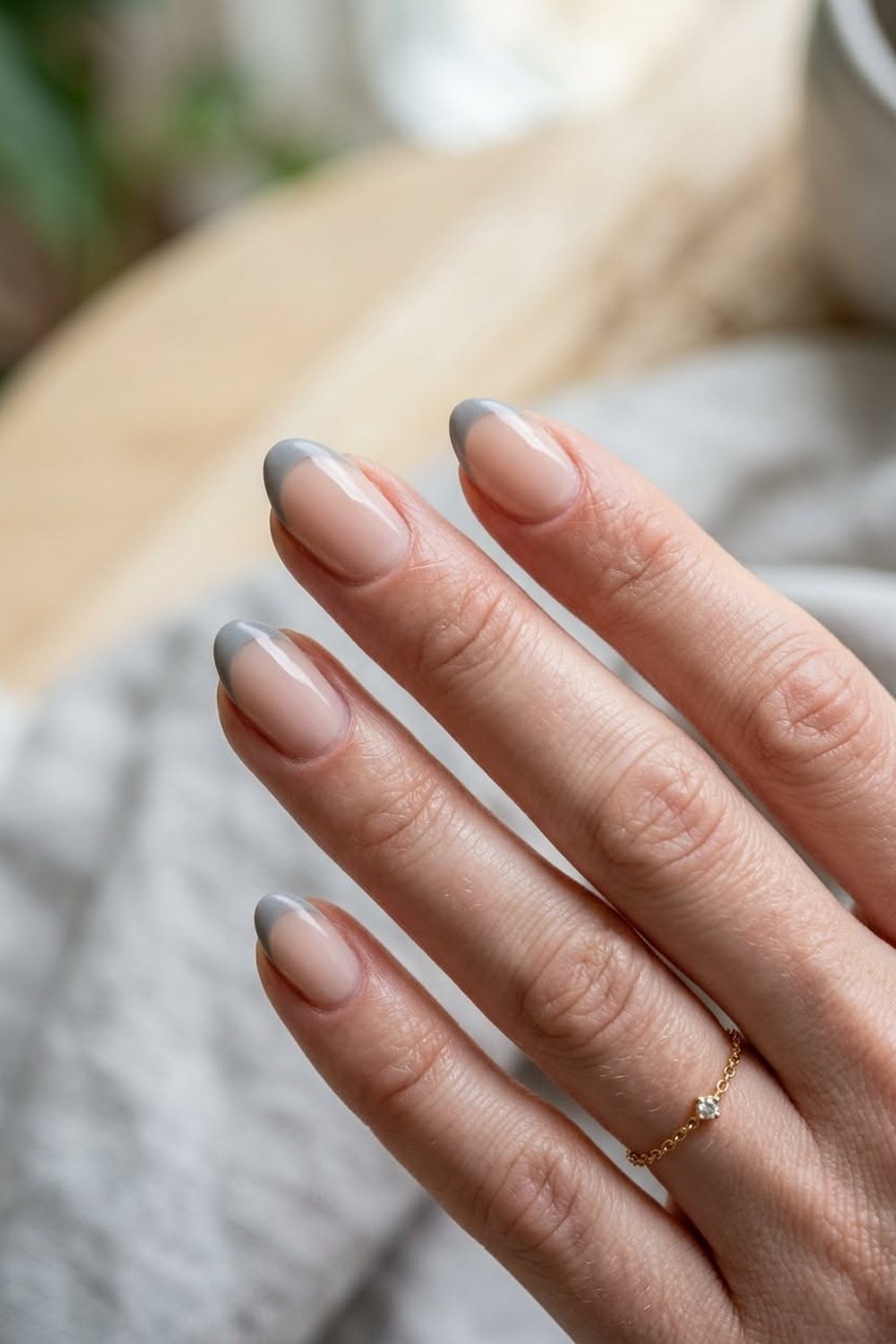 Close-up of a hand with medium almond nails in glossy sheer pink-nude gel with thin soft grey French tips, a fine gold chain ring, against a grey knit and pale wood