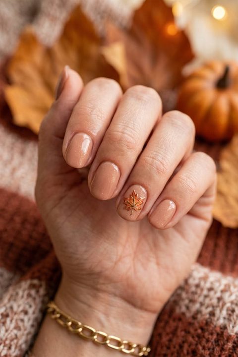 Hand with short squoval sheer peach-nude nails and one tiny orange maple leaf on the ring nail, held in front of autumn leaves and a small pumpkin