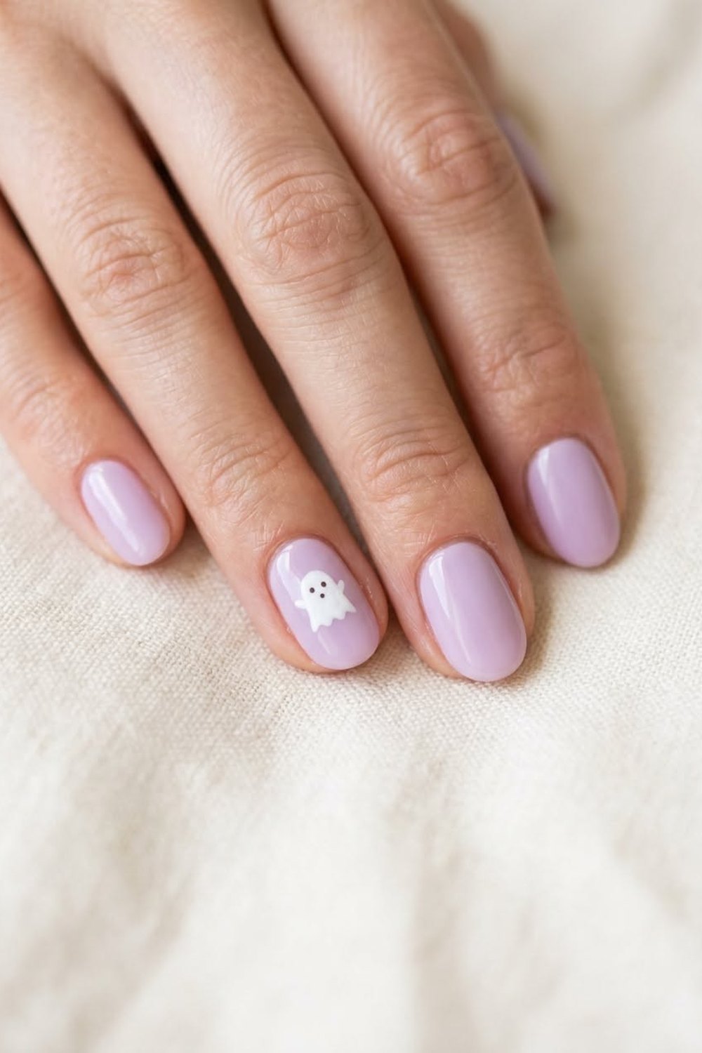 Close-up of a hand with medium oval nails in glossy soft lilac gel and one tiny white friendly ghost on the ring nail, resting on cream linen