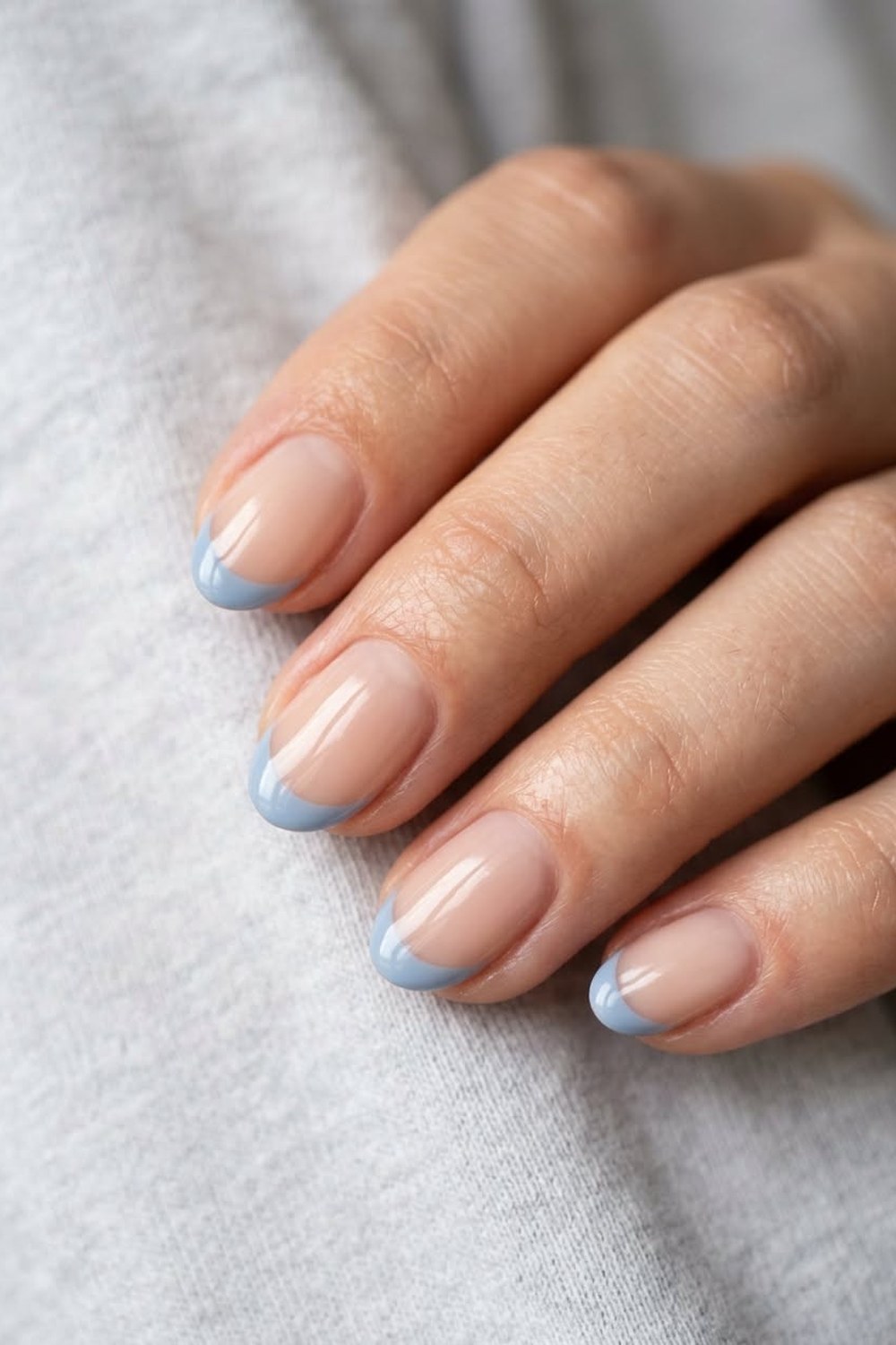 Close-up of a hand with short rounded nails in glossy sheer nude gel with soft powder-blue French tips, resting on a light grey sweater