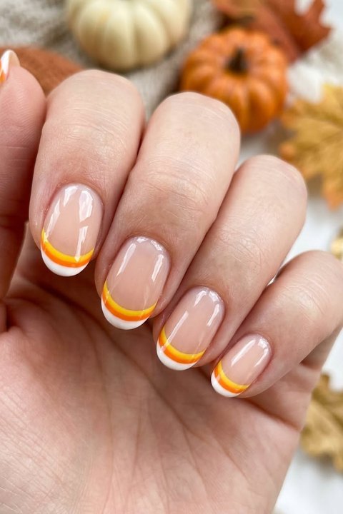 Close-up of a hand with medium almond nails in sheer nude gel and thin white, orange and yellow candy-corn French tips, with small pumpkins and autumn leaves behind