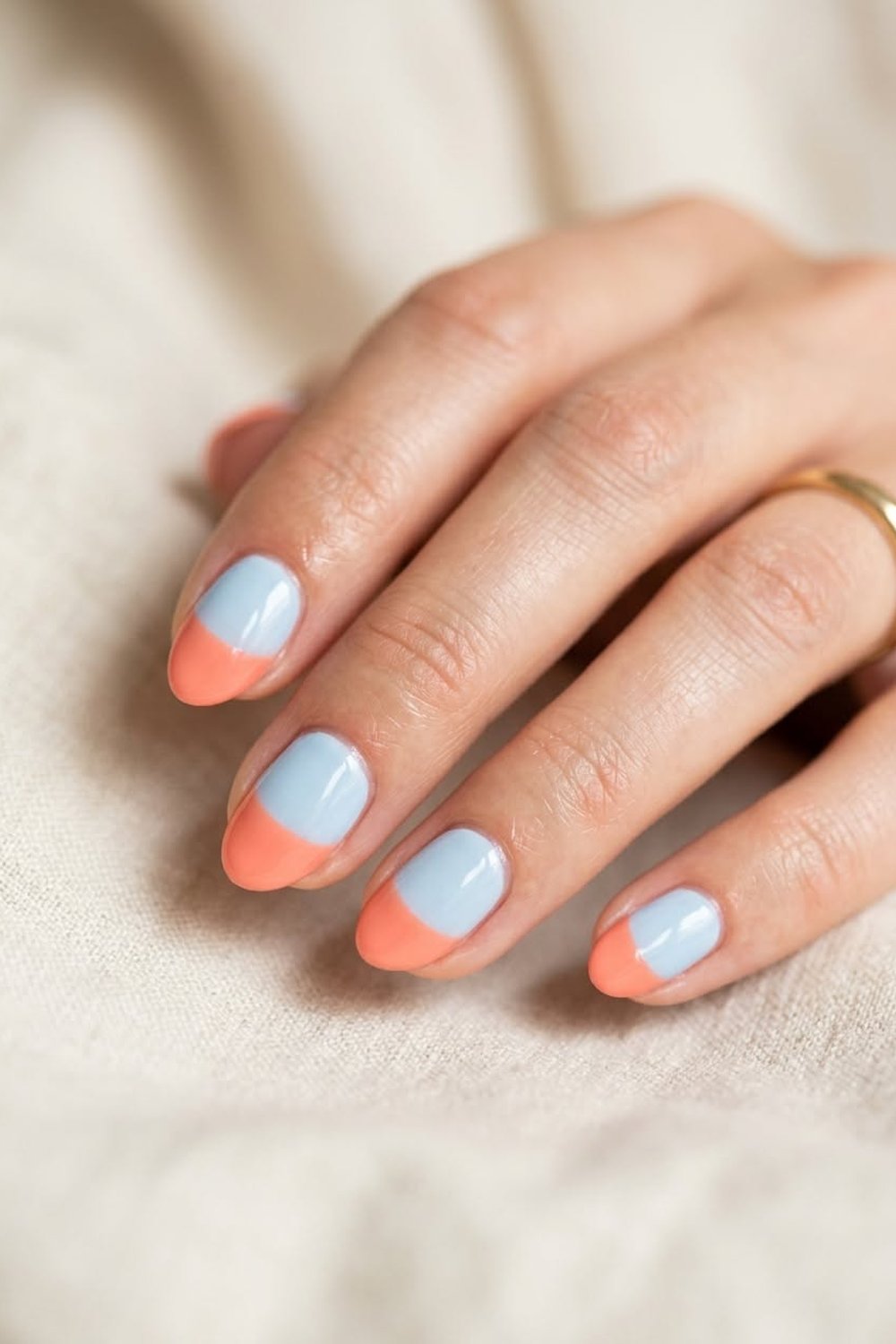 Hand with short rounded nails in soft baby blue with a coral-orange curved tip on each nail, resting on a cream knit