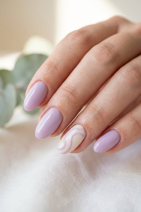 Close-up of a hand with medium almond nails in glossy soft lilac gel and one creamy white accent nail with thin lilac and white swirl lines on the ring finger, resting on white fabric with eucalyptus behind